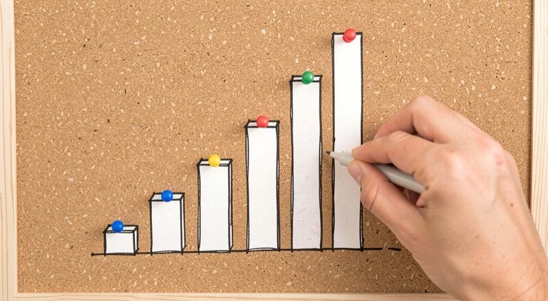 A hand-drawn rising bar chart of plain blocks pinned on a cork board, a hand adding the tallest block, no numbers