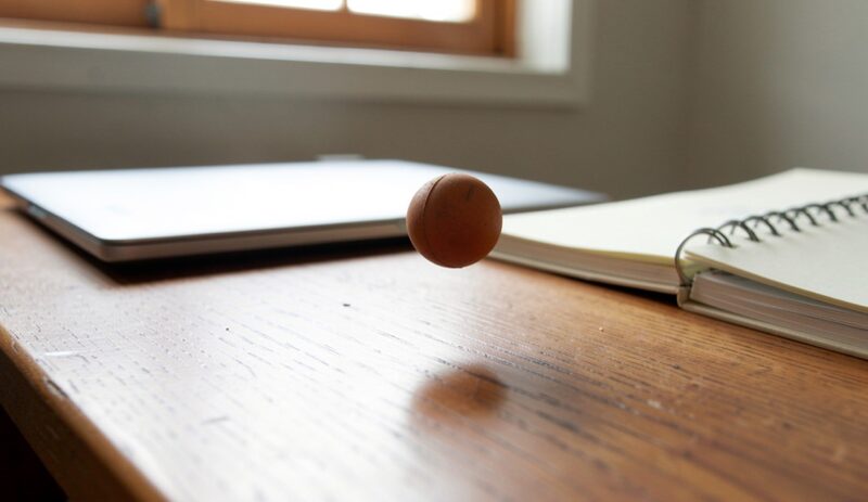 A small rubber ball captured mid-bounce on a wooden desk, a closed laptop and a notebook in the background, window light